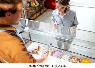 Young Female Client Choosing An Ice Cream Flavor, Standing With A Seller Near The Shop Window