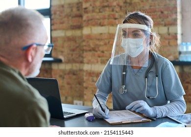 Young Female Clinician In Protective Screen, Mask, And Medical Scrubs Listening To Mature Male Refugee Describing His Symptoms