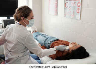 Young Female Clinician In Whitecoat And Protective Mask Scanning Thyroid Area Of Patient Lying On Couch During Ultrasound Examination