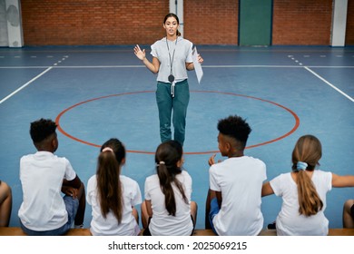 Young Female Coach Talking To Group Of Elementary Students During PE Class At School Gym. 