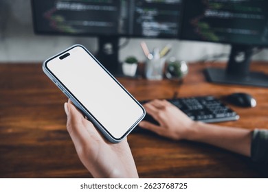 Young female coder holding smartphone with blank screen in modern office workspace featuring coding setup