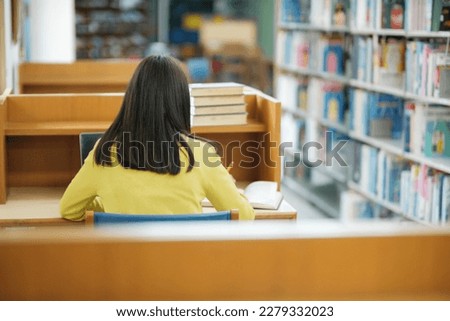 Young female college high school student wearing casual clothings sitting at private desk reading book, studying, writing, and doing research for school project at a library.