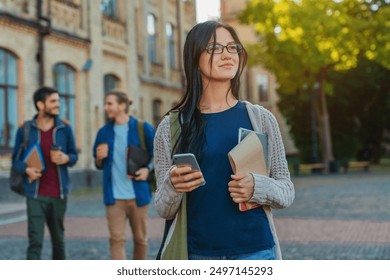 Young female college student woman girl in casual clothes with backpack holding books notebooks using cellphone while standing on university campus look away with classmates students on background - Powered by Shutterstock