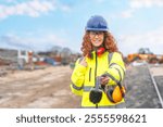 Young female construction worker smiles and gestures while holding safety headphones at a job site