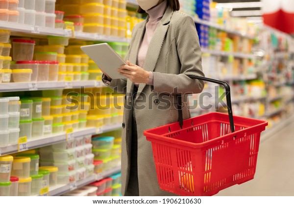 Young female consumer with shopping basket\
standing by large shelf with containers and using touchpad while\
choosing goods in\
supermarket