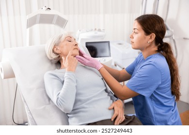 Young female cosmetologist in gloves examining face skin of elderly female client before beauty procedure in spa center indoors - Powered by Shutterstock