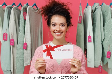 Young female costumer woman wear sweater stand near clothes rack with tag sale in store showroom hold gift certificate coupon voucher card for store isolated on plain pink background studio portrait