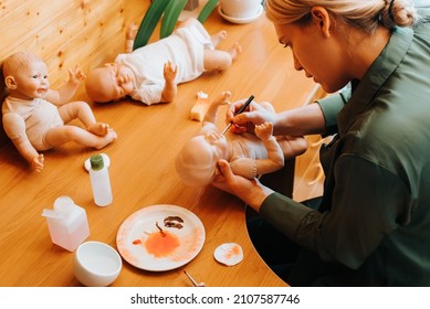 Young female craftsman decorating with brush realistic doll of newborn caucasian child sitting at workplace, indoors. Making handmade dolls, toy craft, hobby reborning concept.