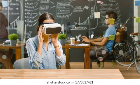 Young Female Creative Developer Wearing Virtual Reality Headset  Works on a Laptop, She Develops New AR, VR Games and Applications. In the Background Stylish Office with Talented People.
