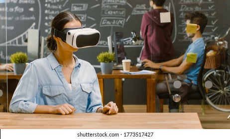 Young Female Creative Developer Wearing Virtual Reality Headset  Works on a Laptop, She Develops New AR, VR Games and Applications. In the Background Stylish Office with Talented People.