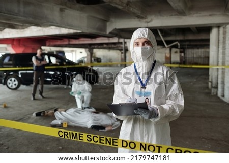Young female crime scene investigator in protective coveralls making notes in document while standing against colleague over dead body