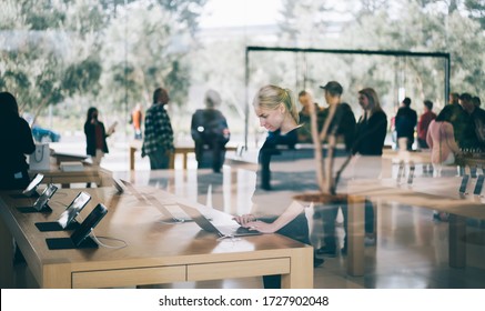 Young Female Customer Standing Near Table With Electronic Products While Examining The Goods, Store Employees And Visitors Stand In Blur On Background. Millennial Hipster Girl Checking Device For Buy