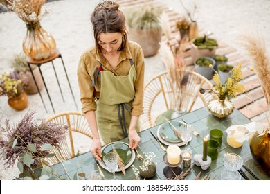 Young female decorator or florist putting herbs on the table, decorating lunch place in natural Boho style in green tones outdoors