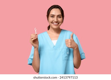 Young female dentist with floss toothpick showing thumb-up on pink background