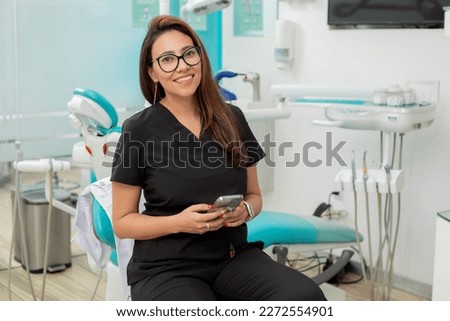 Young female dentist looking at camera smiling, sitting in her office with a cell phone in her hand