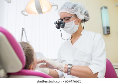 Young Female Dentist Wearing In Loupe Binoculars Checked And Curing Teeth Of Child Patient Sitting In Dental Chair.