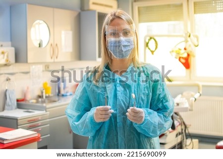 Young female dentist wearing protective clothes, mask on face and face shield, protective gloves holding dental tools standing in office