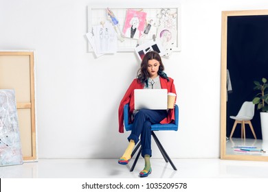 Young female designer working on laptop and holding coffee cup by mood board - Powered by Shutterstock