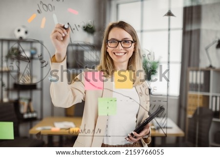 Young female developer smiling and looking at camera while working in creative office environment. Beautiful project manager using digital tablet and office glass board for scheduling project.