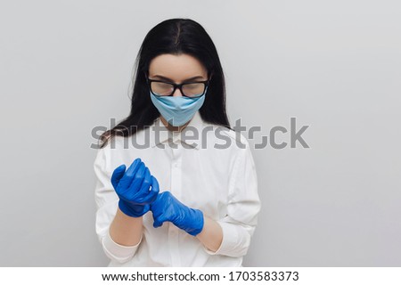 Young female doctor in blue protective medical mask, white smock and glasses wear blue latex gloves before work.The concept of the fight against coronavirus. End of COVID-19. Nursing medical assistant