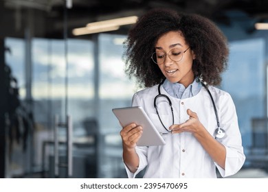 A young female doctor holds a tablet computer in her hands, a woman in a white medical coat consults patients remotely, smiles with satisfaction, stands by the window. - Shutterstock ID 2395470695