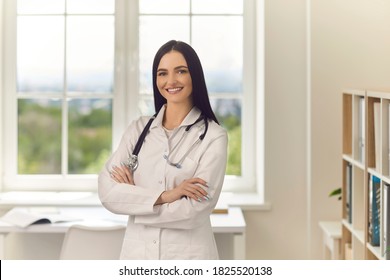 Young Female Doctor In Hospital Office Standing Arms Folded And Looking At Camera Smiling. Friendly Clinic Staff Member At Work. Portrait Of Modern General Practitioner, Physician, Healthcare Provider