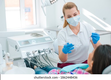 Young female doctor in medical mask and rubber gloves sitting near the patient on a dental chair and holding mouth mirrors