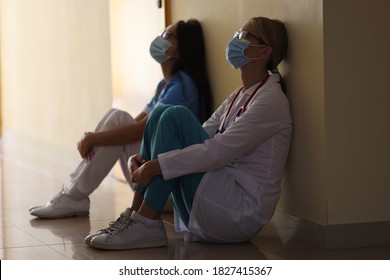 Young Female Doctor And Nurse In Medical Masks Sit In Corridor Of Hospital. Emotional Burnout Of Healthcare Workers During Covid19 Pandemic Concept.