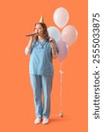 Young female doctor in party hat with balloons, whistle and birthday cupcake on orange background