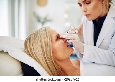 Young Female Doctor Performing A Botox Injection To The Forehead Of A Mature Woman Lying On A Table In A Beauty Clinic