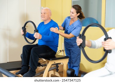 Young female doctor of rehabilitation center helping elderly man to do remedial gymnastic with pilates resistance ring - Powered by Shutterstock