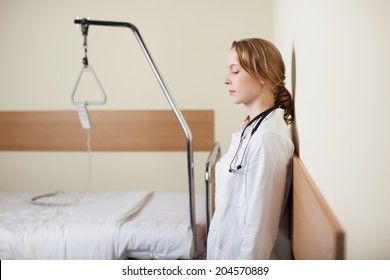 Young Female Doctor Standing With Closed Eyes Leaning Against The Wall In A Ward Alongside An Empty Hospital Bed As She Takes A Moment To Gather Herself Together