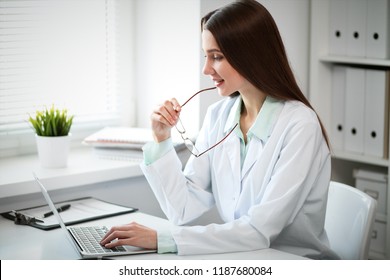 Young female doctor typing on laptop computer while sitting at the table near the window in hospital office - Shutterstock ID 1187680084