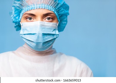 young female doctor in uniform, medical cap and face mask, close-up portrait. with a serious gaze looking at the camera on a blue background. copy space - Powered by Shutterstock