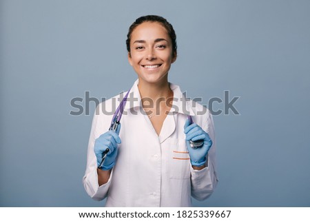 Young female doctor wearing latex gloves and white medical coat holding stethoscope on blue background.