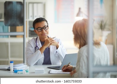 Young female doctor in white coat sitting at the table and listening to patient sitting in front of her at office