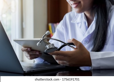 Young female doctor in white lab coat, hand holding medical stethoscope and using modern digital tablet pc, work on laptop computer at office. Online medical, e health or medical network concept. 