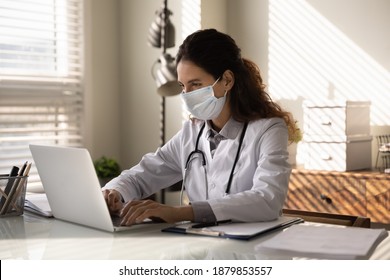 Young Female Doctor In White Medical Uniform And Facial Mask Look At Laptop Screen Consult Patient Online. Woman GP Or Physician In Facemask Have Webcam Consultation, Work On Computer In Clinic.