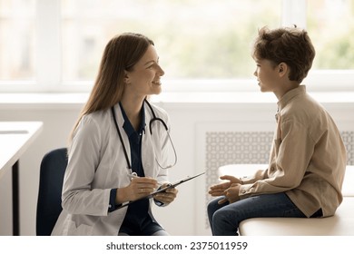 Young female doctor in white uniform hold clipboard listen boy at checkup meeting, write symptoms, make health test, ask about illness. Little patient visited clinic get consultation from pediatrician - Powered by Shutterstock