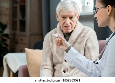 Young female doctor in whitecoat looking at thermometer after measuring body temperature of sick senior man sitting on couch in living-room - Shutterstock ID 1859283091