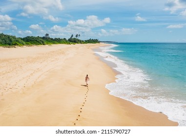 Young female dressed in light summer clothes walking barefoot leaving footprints on the sand on Indian ocean Tangalle lonely coconut trees beach on Sri Lanka island. Aerial top view drone shot.