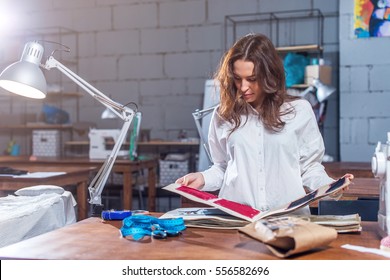 Young female dressmaker choosing material from catalogue in studio. Tailor looking through fabrics while standing in a sewing workshop