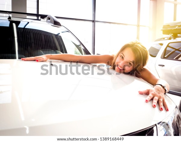 Young female driver embracing hood of new\
car. Dream about car. Gorgeous smiling woman hugging lies on the\
hood of new red car in the\
dealership.