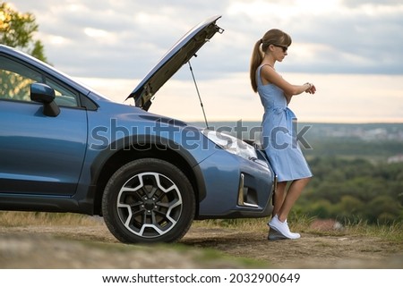 Young female driver standing near a broken car with open up hood inspecting her vehicle engine and waiting for help.