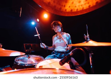 Young female drummer energetically playing drums on a stage illuminated by vibrant, colorful lighting, creating a dynamic and captivating musical performance - Powered by Shutterstock