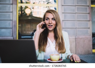 Young female in elegant clothes looking amazed while talking on mobile phone and sitting with laptop and cup of hot drink in outdoor cafe - Shutterstock ID 1728283765