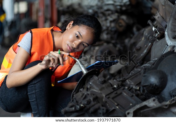 Young female employee warehouse worker using\
clipboard checking old automotive spare parts, engine, motor,\
machine at the garage industry factory or automotive spare parts\
storage warehouse