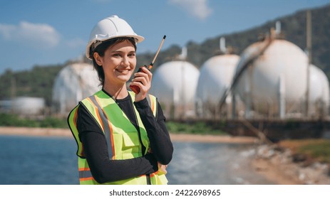 A young female engineer wearing a safety helmet and reflective vest holds a walkie-talkie at an industrial storage facility. - Powered by Shutterstock