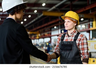 Young female engineer in workwear looking at factory manager during handshake while both standing in front of each other