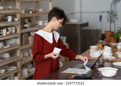 Young Female Entrepreneur Looking At Bills, Tracking Expenses, Craftswoman Potter Studio Owner Doing Bookkeeping, Paying Income Taxes While Working In Ceramic Shop. Entrepreneurship And Small Business